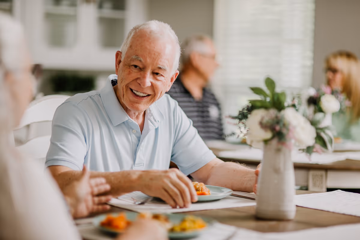 An elderly man with white hair wearing a light blue polo shirt is smiling and engaging in conversation while sitting at a dining table with a plate of food. In the background, other elderly individuals are seated at tables in a bright, well-lit room with white walls and windows. A vase with flowers is placed on the table in the foreground.