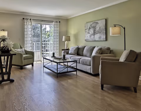 A bright living room with light green walls, a large gray sofa with cushions, two armchairs, a black metal and glass coffee table, a floor lamp, a table lamp, and a sliding glass door leading to a balcony with a view of trees outside.