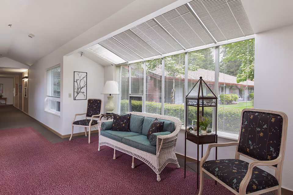A bright indoor sitting area with a wicker sofa with green cushions and two floral upholstered chairs. There is a small table with a lamp and a decorative glass terrarium. Large windows with blinds allow natural light and show a view of greenery and buildings outside. The floor is covered with a patterned red carpet.