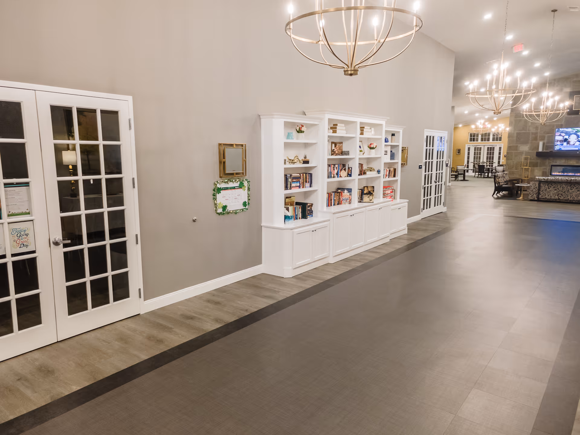 Spacious, well-lit interior common area with white bookshelves along a wall, chandeliers overhead, and seating with a fireplace visible in the distance.