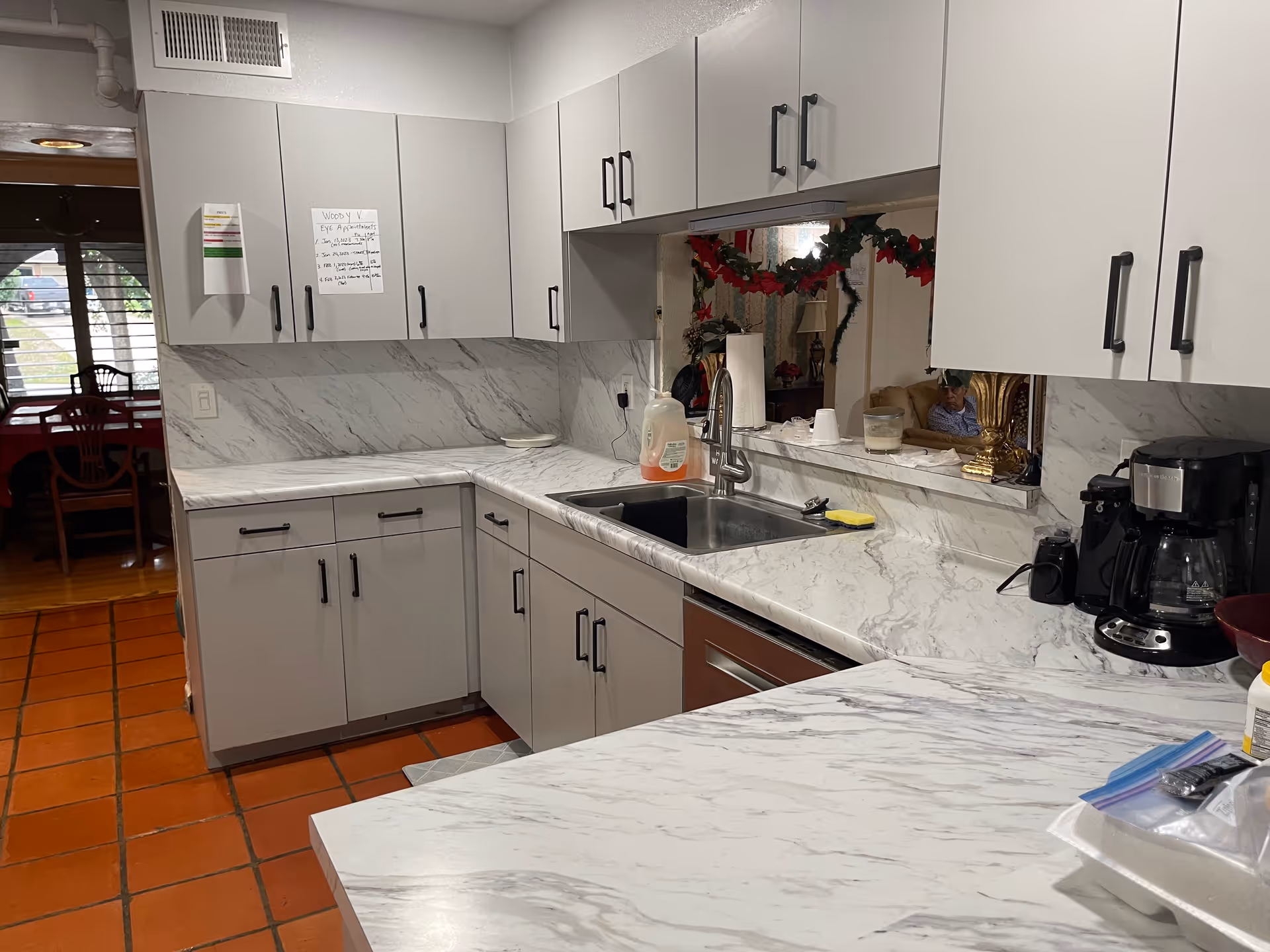 A kitchen area with white marble countertops and backsplash, light gray cabinets with black handles, a stainless steel sink, a coffee maker, and various kitchen items. There is a pass-through window decorated with holiday garland, showing a living room area with a person sitting on a couch. The floor is covered with reddish-brown tiles, and a dining area with wooden chairs is visible through an open doorway.