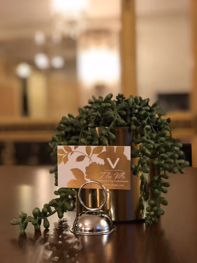 A close-up of a small green succulent plant in a metallic pot on a wooden table. In front of the pot is a silver bell and a business card holder displaying a card with the logo and name 'The Villa Senior Living Community'. The background is softly blurred, showing warm indoor lighting and furniture.