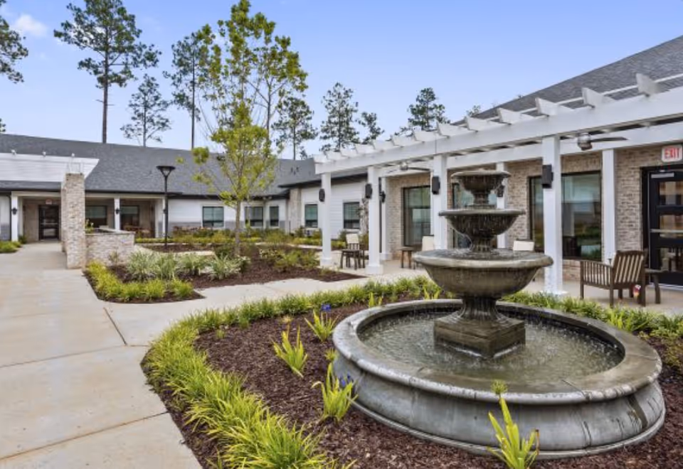 Outdoor courtyard with a tiered fountain, landscaped beds, walkways and seating under a pergola in front of a single-story building.