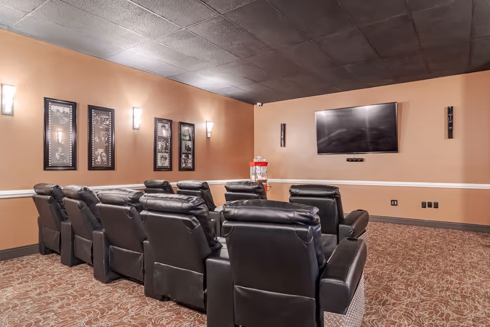 A cozy home theater room with two rows of black leather recliner chairs facing a large flat-screen TV mounted on a beige wall. The room has patterned carpet flooring, wall sconces providing soft lighting, and framed black-and-white photos on one wall. A popcorn machine is placed in the corner near the TV.