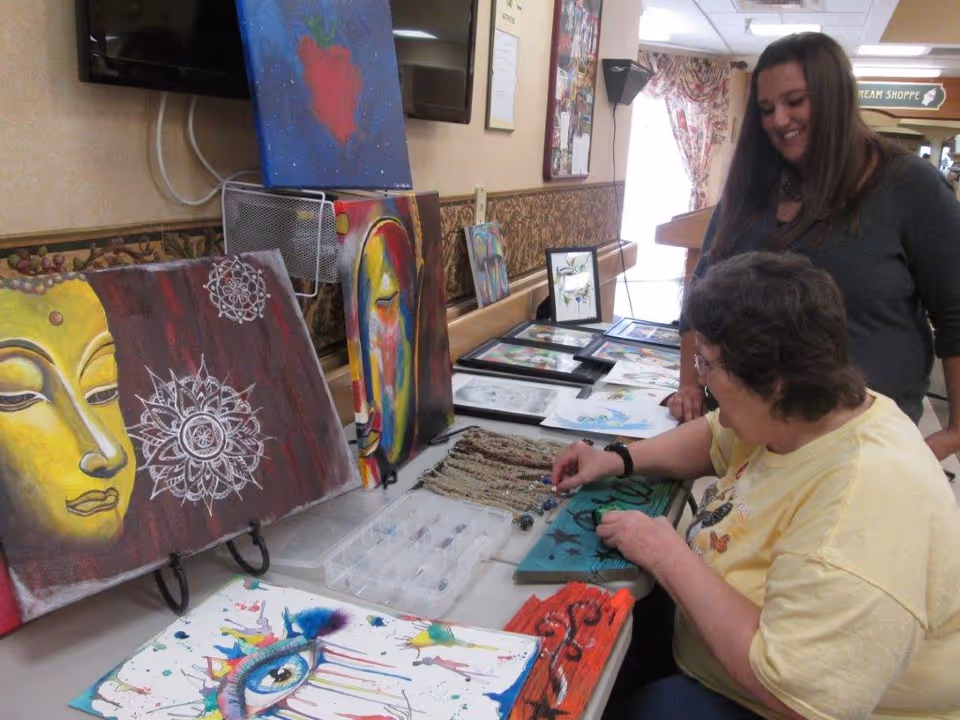 Two women engaged in an art activity at a table displaying various colorful paintings and artwork. One woman is seated and working on a craft project, while the other stands nearby smiling. The setting appears to be an indoor common area with patterned wallpaper and a window with floral curtains.