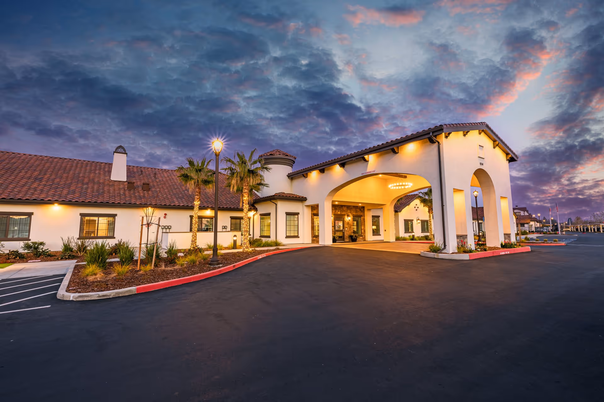 Exterior view of The Gardens At Laguna Springs facility at dusk, showing a large entrance with arches, warm lighting, palm trees, and a tiled roof under a dramatic cloudy sky.