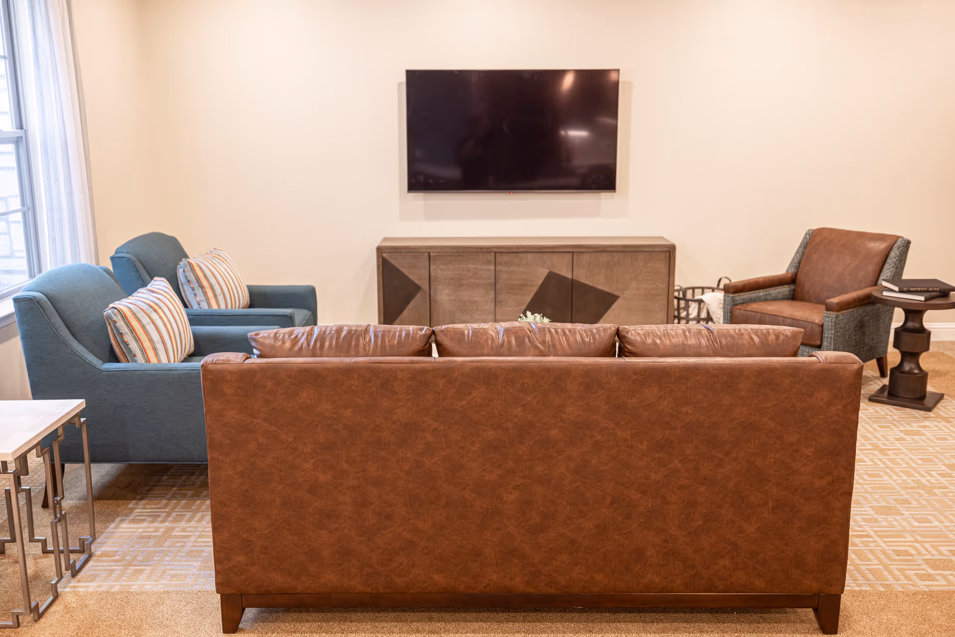 A cozy living room with a brown leather sofa facing a wall-mounted flat-screen TV. To the left, there are two blue armchairs with striped cushions, and to the right, a single armchair with a brown leather seat and a small side table next to it. The room has a beige patterned carpet and a wooden cabinet beneath the TV.
