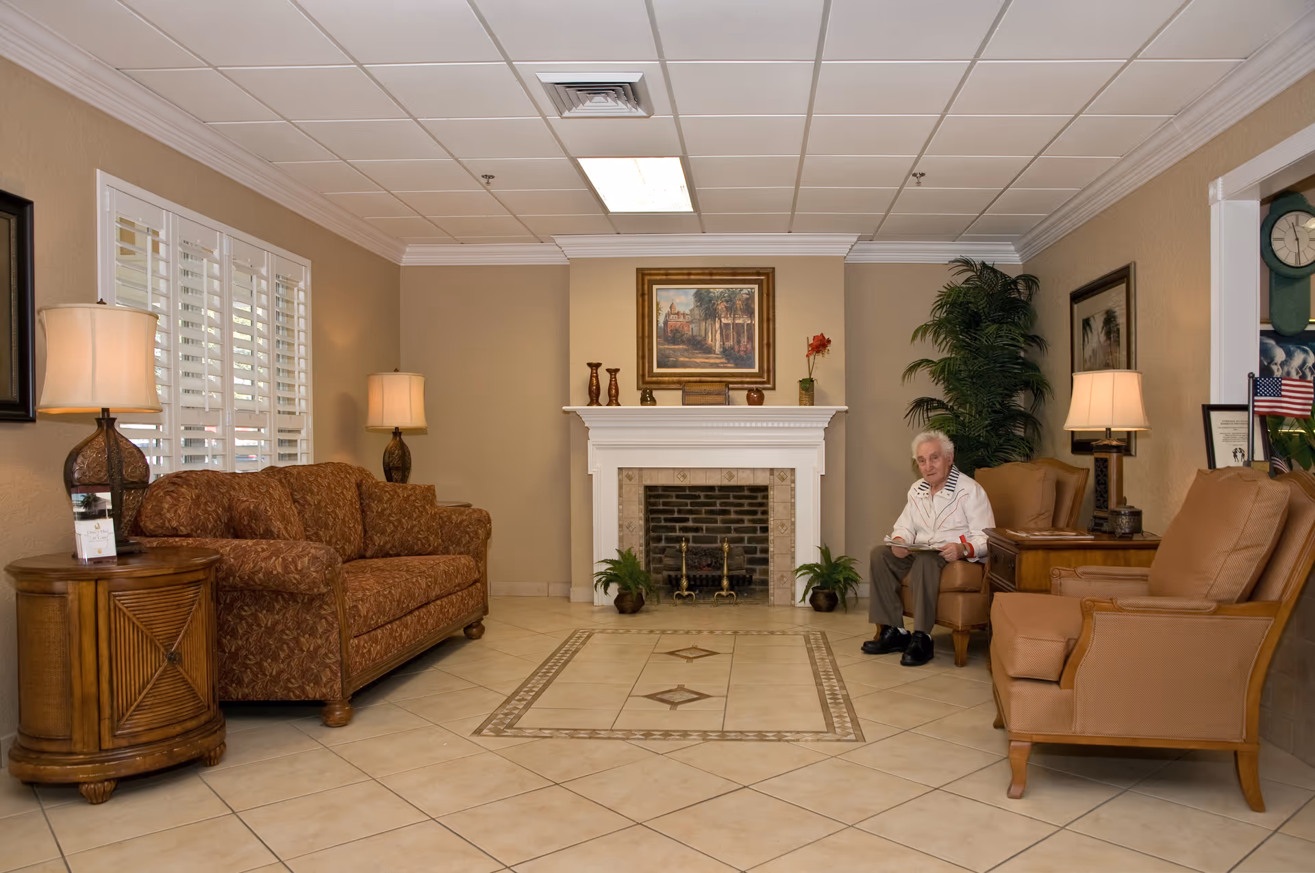 A comfortable senior living lounge with sofas, armchairs, a fireplace and an elderly woman seated in a chair.