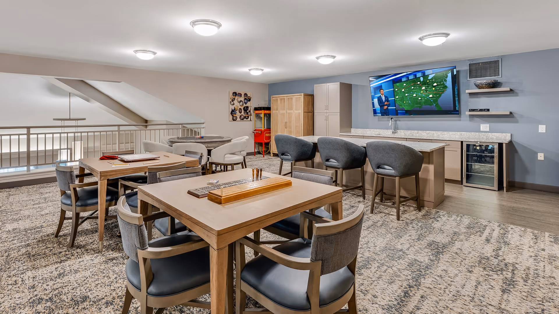 A spacious common area with multiple tables and chairs arranged for games and socializing. There is a countertop with bar stools, a large flat-screen TV mounted on the wall showing a weather forecast, and a popcorn machine in the corner. The room has a modern design with carpeted flooring and overhead lighting.