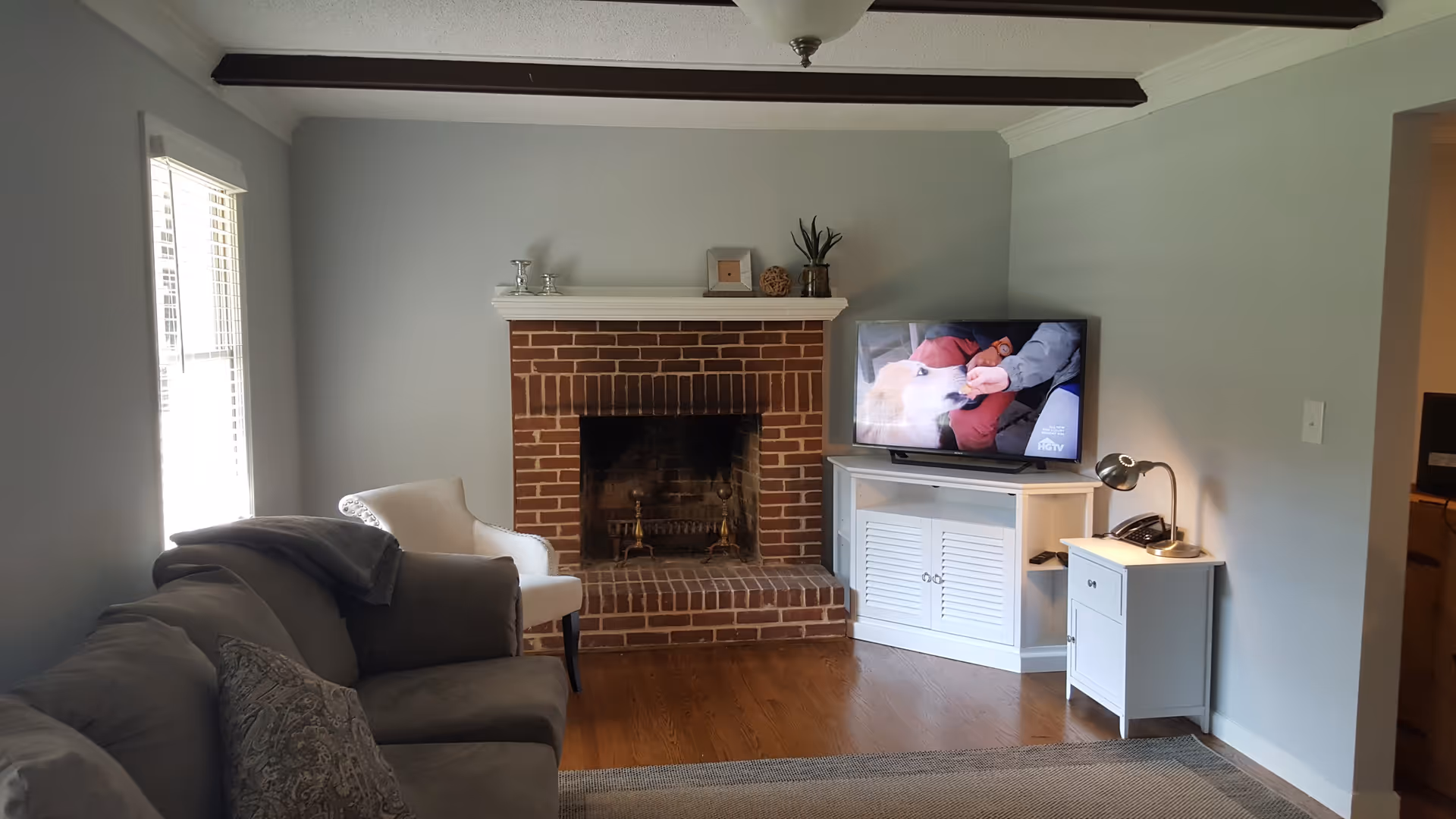 Living room with a brick fireplace, TV on a white stand, a sofa and armchair, and hardwood floors.
