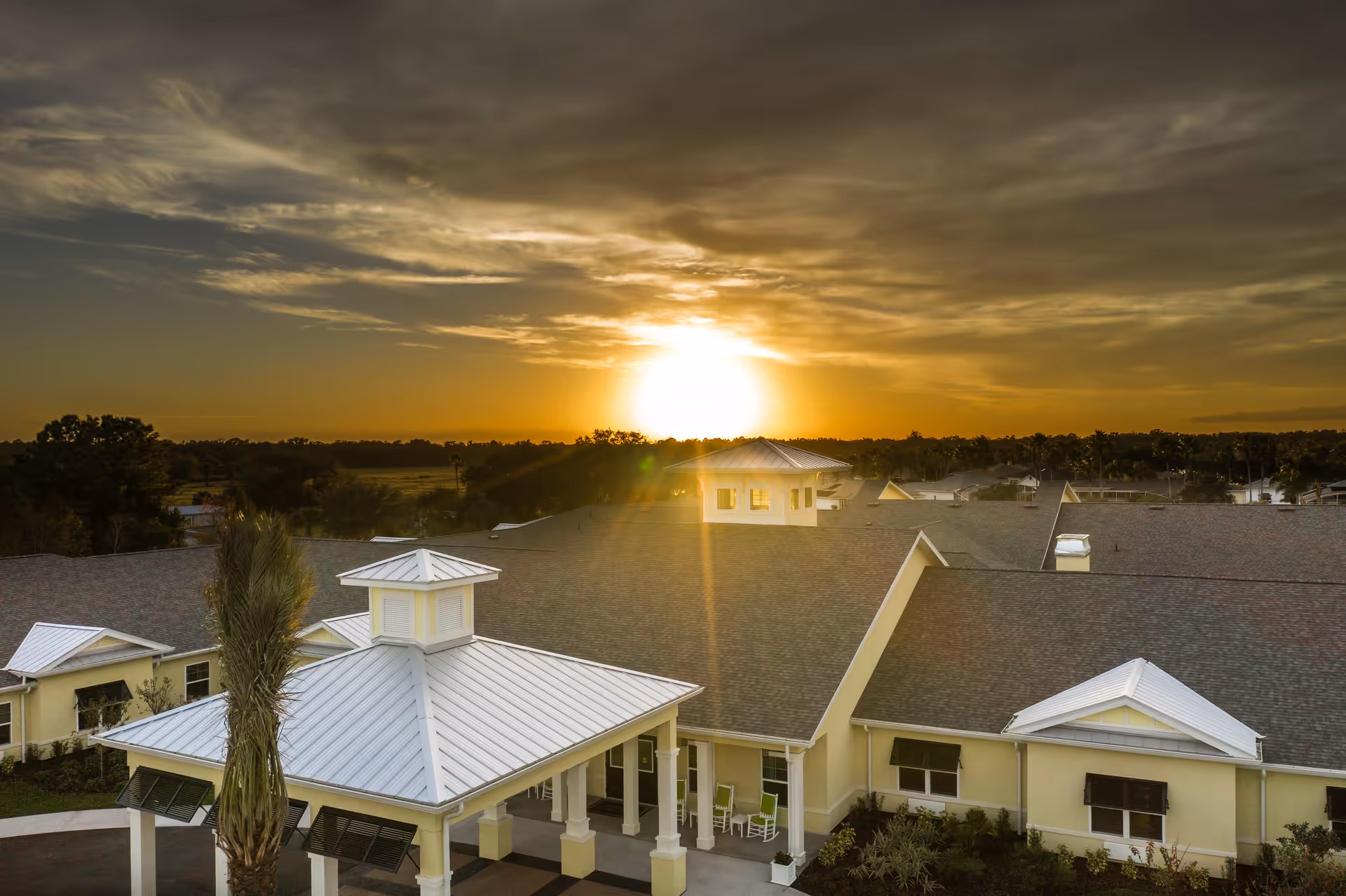 Exterior view of The Addison of Port Orange senior living facility at sunset, showing the building's roof, entrance with white pillars, and surrounding landscape under a partly cloudy sky.