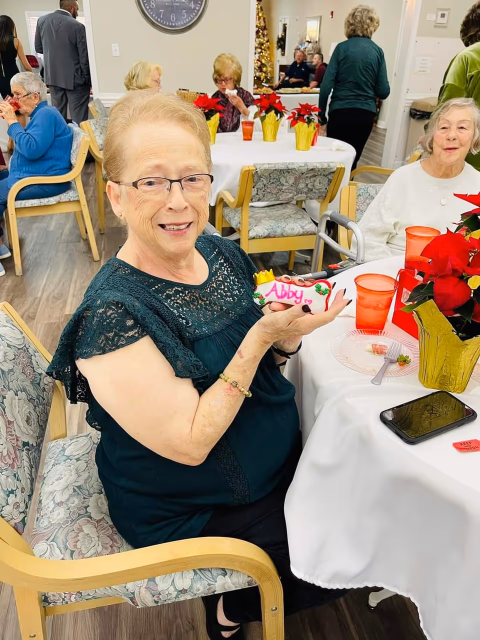 An elderly woman wearing glasses and a dark green lace top is sitting at a table in a senior living community dining area, smiling and holding a decorated cookie with the name 'Abby' written on it. The table is covered with a white tablecloth and has red poinsettia plants in gold foil pots, red plastic cups, and a smartphone. Other elderly residents are seated and standing in the background, with festive holiday decorations visible.
