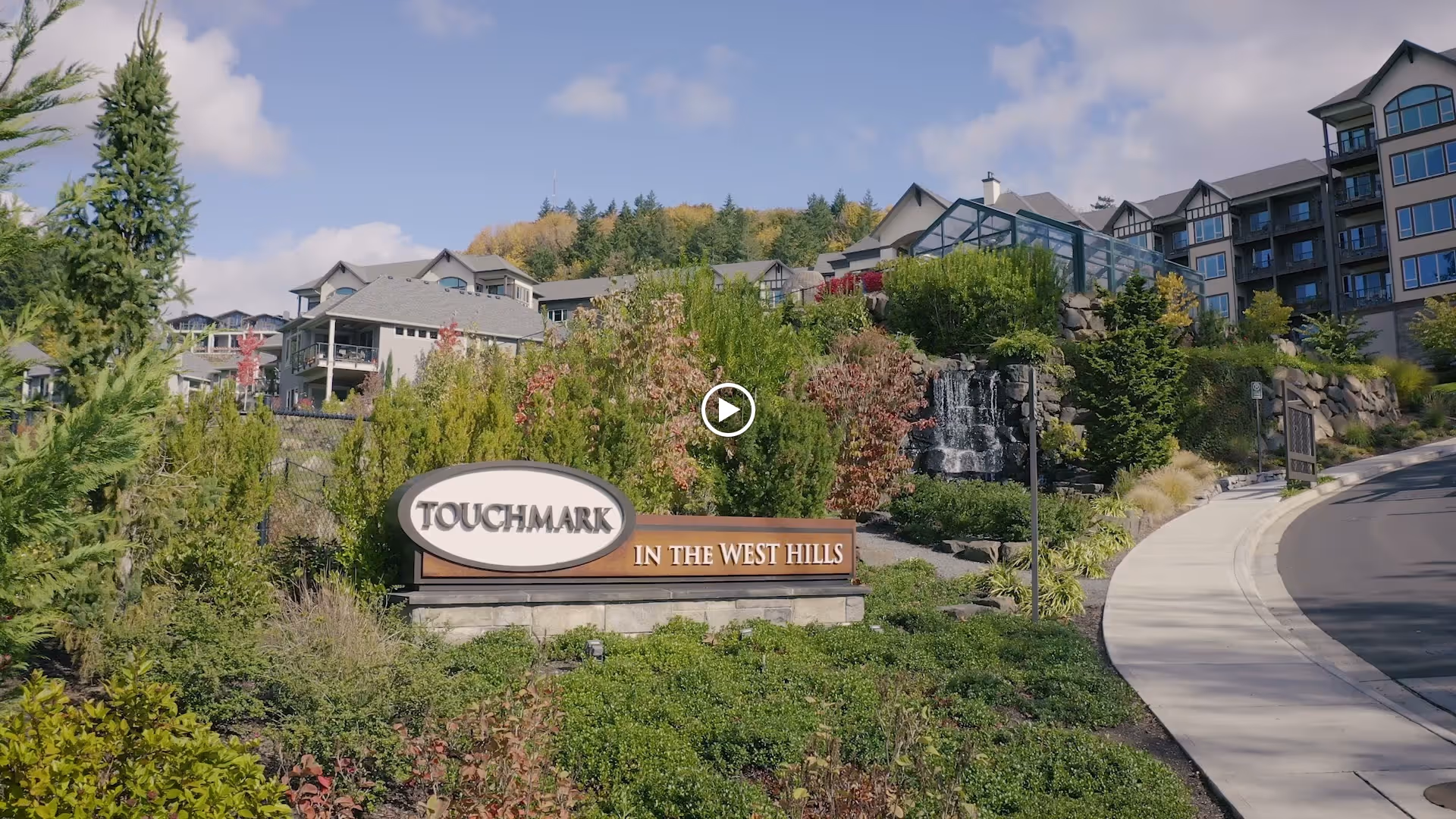 Entrance sign for Touchmark in the West Hills surrounded by greenery and landscaping, with a large multi-story residential building in the background under a partly cloudy sky.