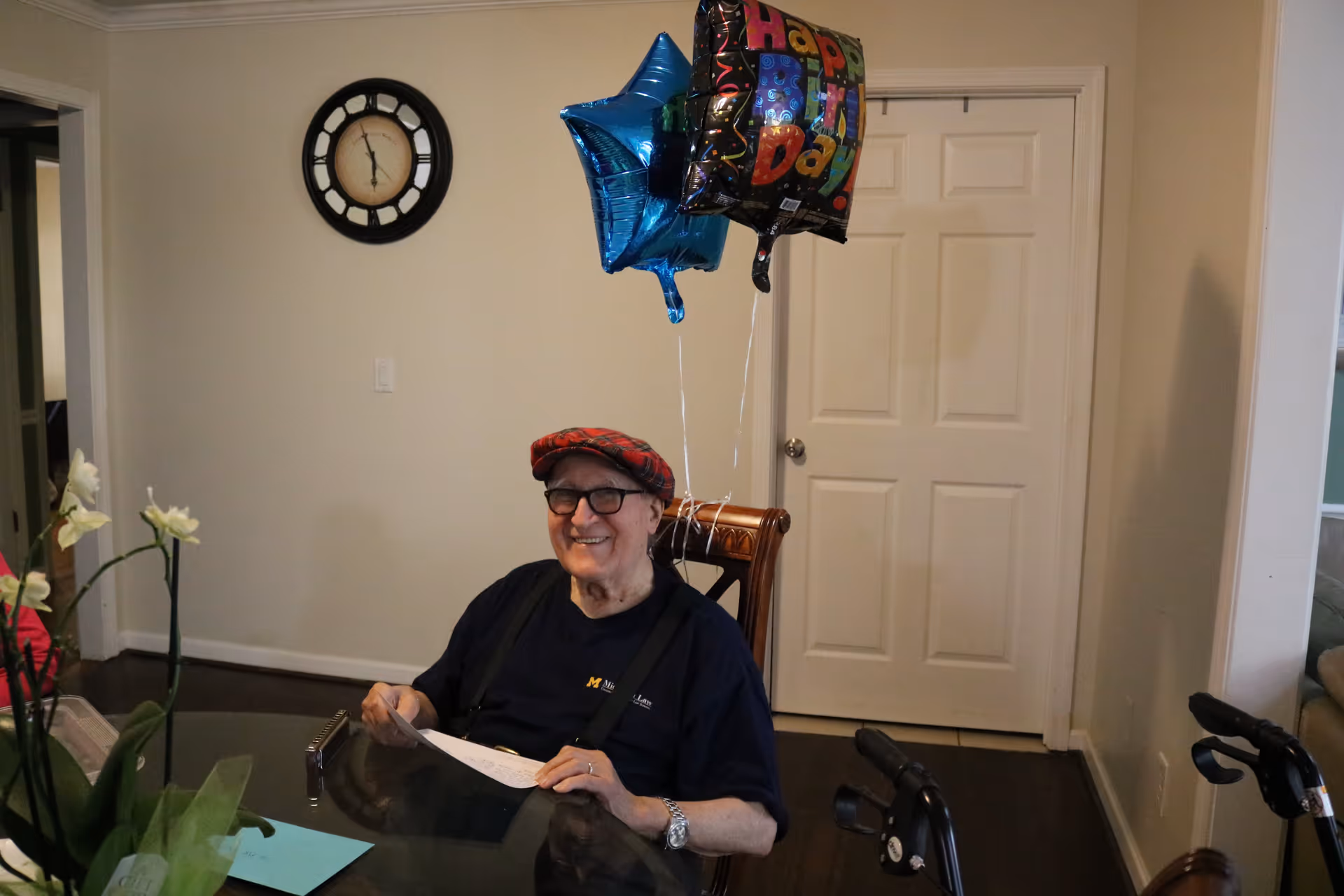 An elderly man wearing glasses and a red plaid hat is sitting at a dining table with a smile. Behind him are two balloons, one blue star-shaped and one with a colorful 'Happy Birthday' message. A clock is mounted on the wall, and a walker is visible near the table.