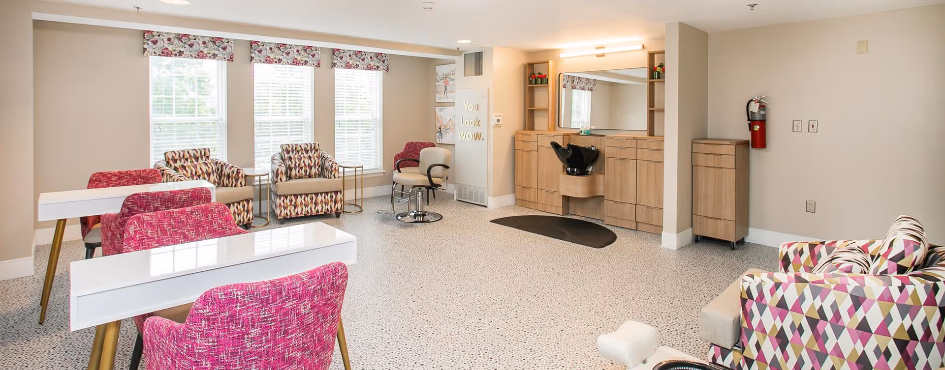 A bright and spacious salon area with multiple styling chairs and mirrors. The room features large windows with floral valances, patterned armchairs, white manicure tables with pink chairs, and a hair washing station. The walls are painted light beige, and there is a fire extinguisher mounted on one wall.