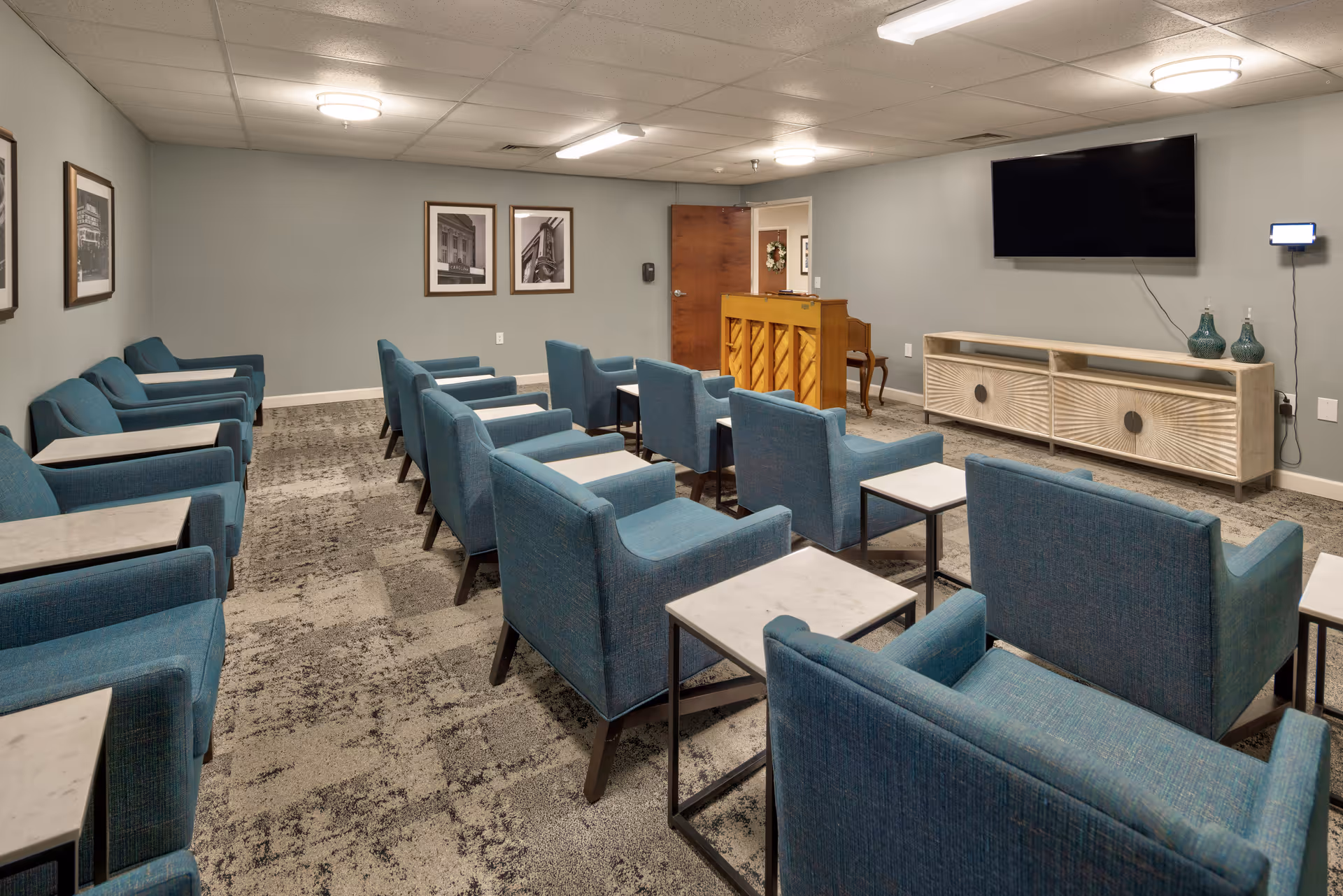A small auditorium or meeting room with rows of blue upholstered chairs each paired with a small white side table. At the front of the room is a wooden piano, a large flat-screen TV mounted on the wall, and a light-colored media console with decorative vases. The walls are painted light gray and decorated with framed black and white photographs. The ceiling has recessed lighting and a few round ceiling lights.