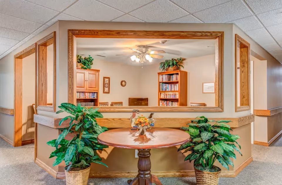 Interior view of a senior living facility showing a round wooden table with a floral centerpiece and two potted plants on either side. Behind the table is a large window opening into a room with bookshelves filled with books, a ceiling fan with lights, and framed pictures on the walls.