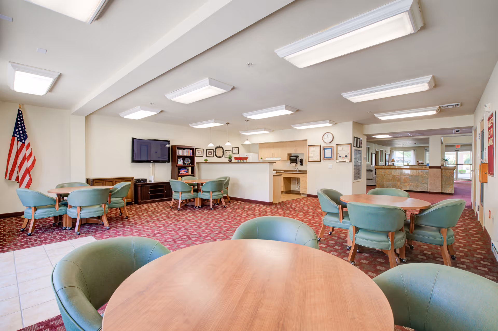 Bright communal lounge/dining area with round wooden tables, green upholstered chairs, a wall-mounted TV, and a reception counter.