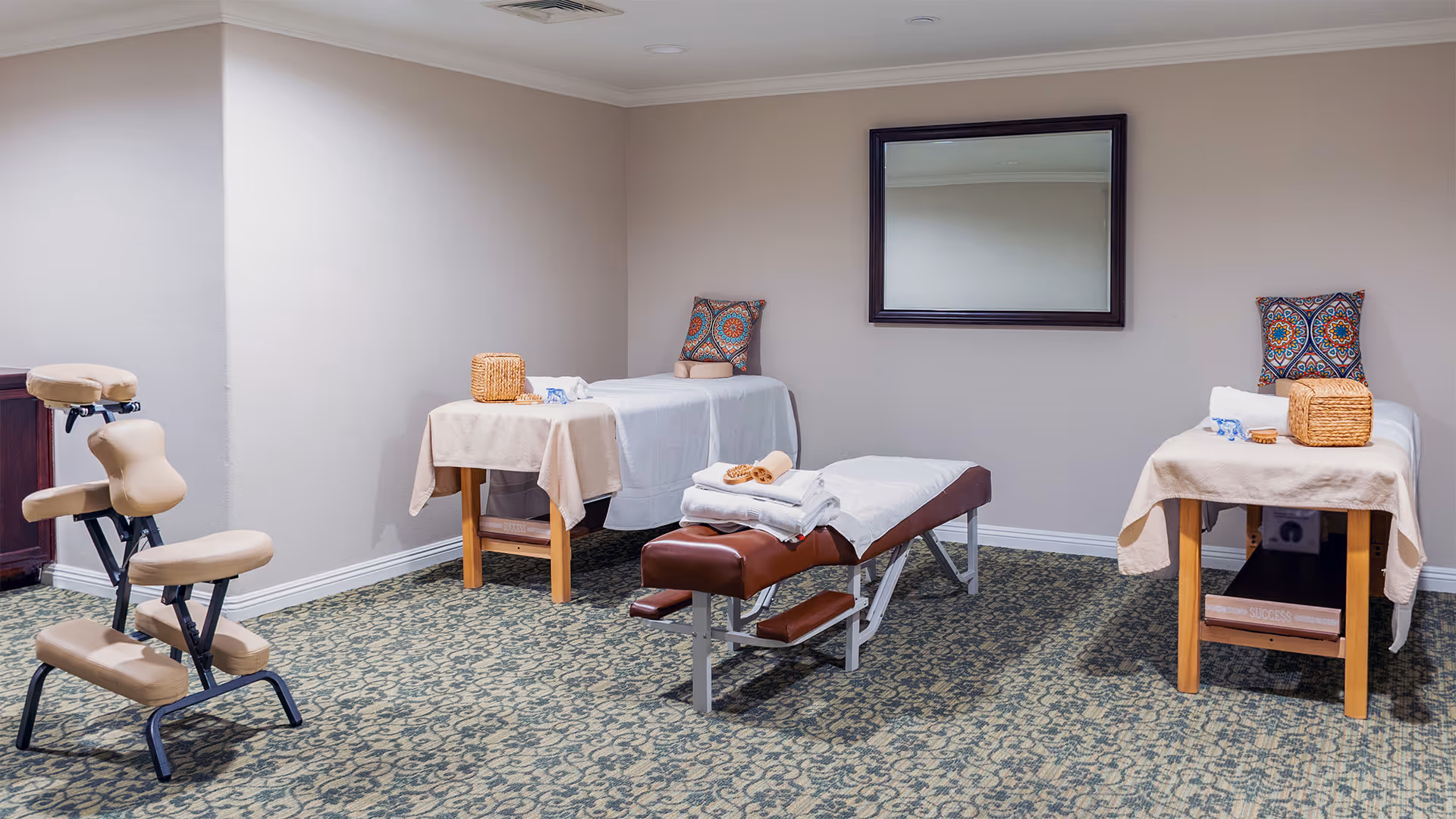 A calm therapy room with three treatment tables, a massage chair, folded towels, and a wall mirror.