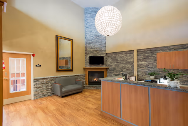 Reception area of a senior living facility with a wooden front desk, a gray couch, a wall-mounted TV above a fireplace, stone accent walls, a large round pendant light, and a door with glass panels.