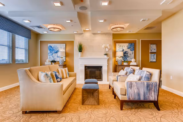A bright and elegant living room with two beige sofas and two armchairs arranged around a small wooden coffee table. The room features a white fireplace centered on the far wall, flanked by decorative blue floral artwork and vases. Large windows with blinds allow natural light to fill the space, and the ceiling has recessed lighting and modern light fixtures.