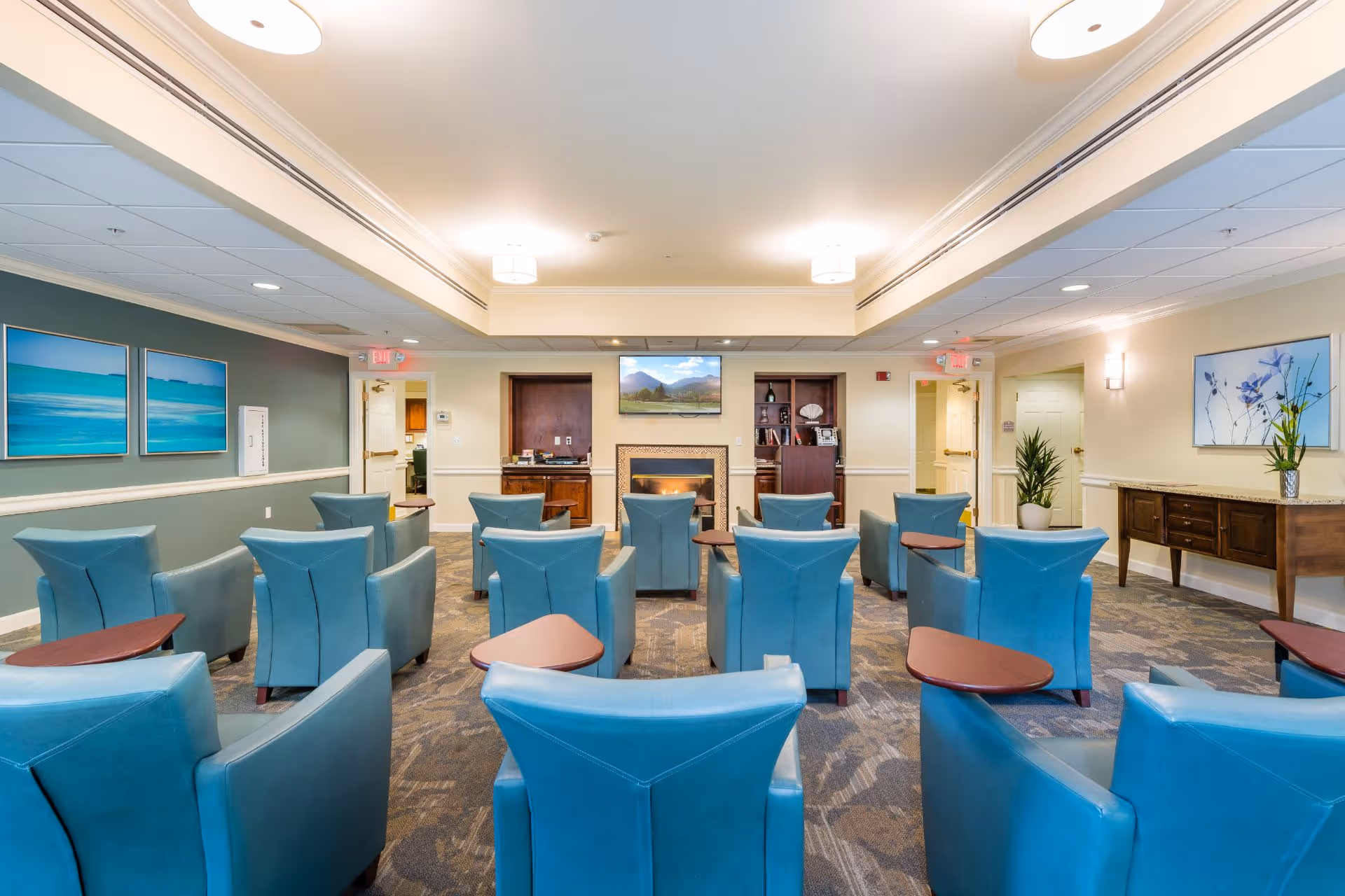 A well-lit common area with multiple blue armchairs arranged in rows facing a wall-mounted television above a fireplace. The room has carpeted flooring, light-colored walls, and decorative paintings on the walls. There are side tables attached to some chairs and a wooden console table with a plant on the right side.