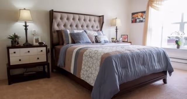 A neatly made bed with a tufted headboard in a bedroom. The bed has a striped comforter in shades of blue, gray, and beige, with multiple pillows. On either side of the bed are nightstands with lamps, a small plant, and framed pictures. A window with sheer curtains lets in natural light, and a small orchid plant is on the windowsill.
