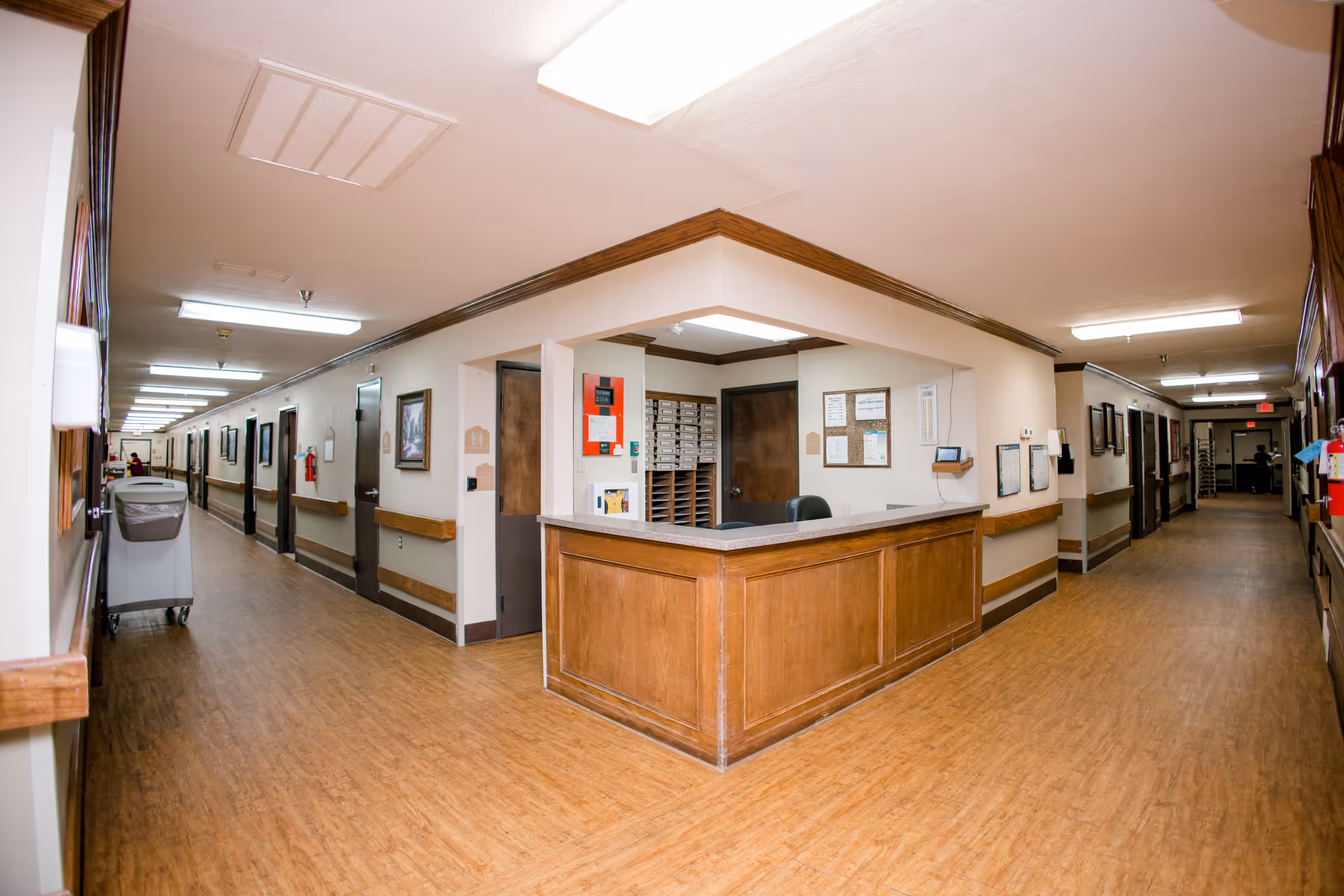 Interior view of a nursing facility hallway with wooden flooring and beige walls. A wooden reception desk is positioned at the center with mail slots and bulletin boards behind it. The hallway extends to the left and right with several closed doors, handrails along the walls, and fluorescent ceiling lights.