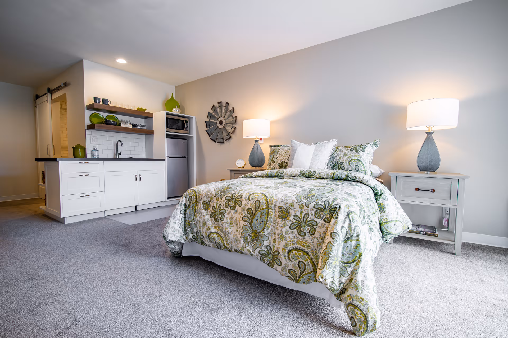 A cozy bedroom with a bed covered in a green and white patterned comforter and pillows. On either side of the bed are matching nightstands with lamps. To the left, there is a small kitchenette area with white cabinets, a sink, a microwave, and a refrigerator. The room has light gray carpet and neutral-colored walls with a decorative wall clock above the nightstand.