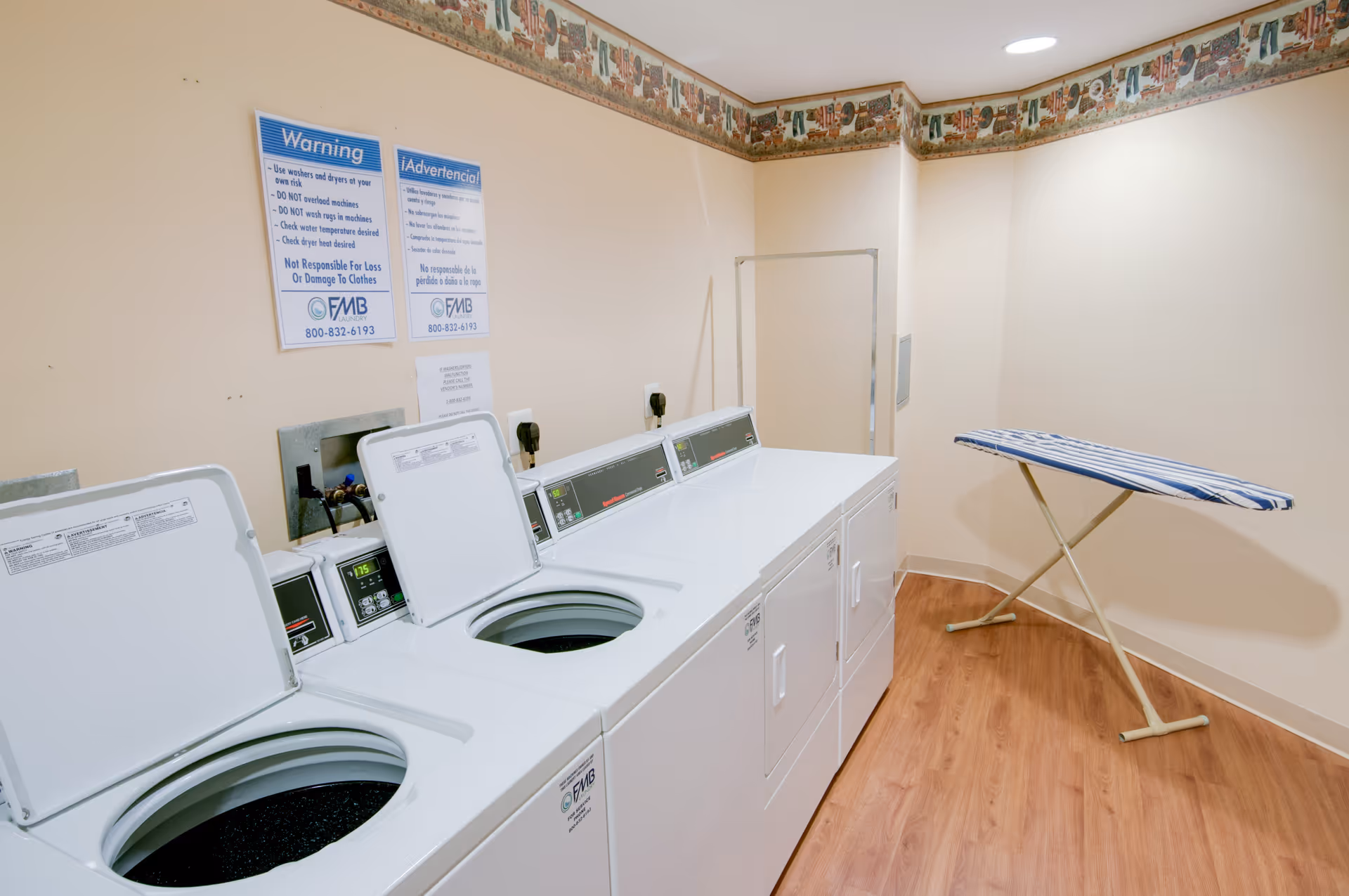 Laundry room with three top-loading washing machines and two front-loading dryers. An ironing board with a striped cover is set up against the wall. The room has light-colored walls with a decorative border near the ceiling and wood-style flooring.