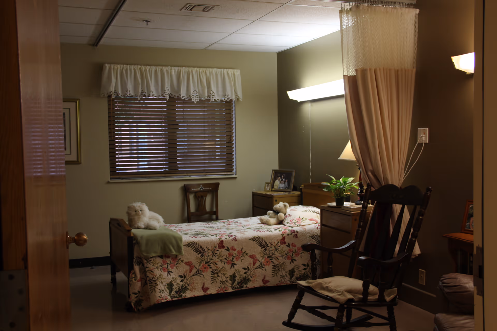 A cozy bedroom in a senior living facility with a single bed covered in a floral quilt, two stuffed animals on the bed, a wooden rocking chair with a cushion, a small wooden nightstand with a potted plant and framed photos, a window with blinds and a white valance, and a privacy curtain partially drawn.
