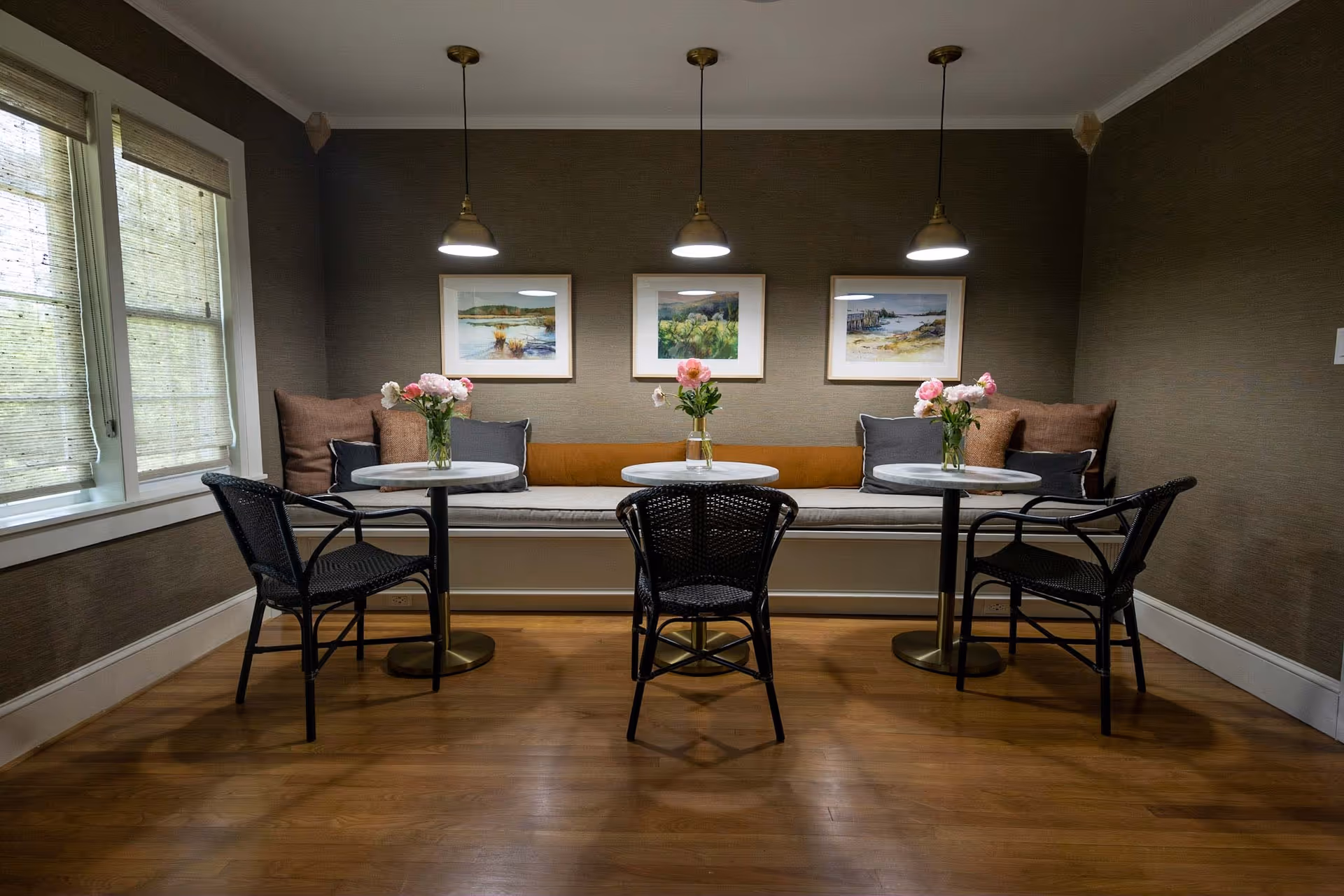 Cozy dining nook with a built-in bench, three pendant lights, round tables with vases of flowers and wicker chairs.