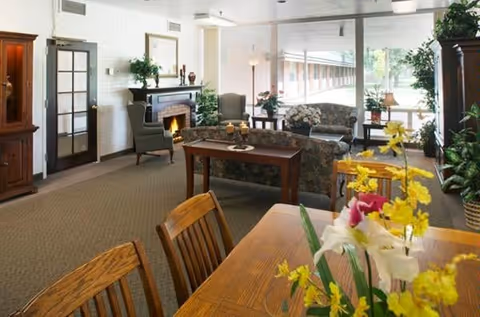A cozy living room area in a senior living facility featuring wooden chairs and a table with a floral centerpiece in the foreground. The room includes a fireplace with a mirror above it, two armchairs, a floral-patterned sofa, and large windows letting in natural light. There are various plants and lamps decorating the space.