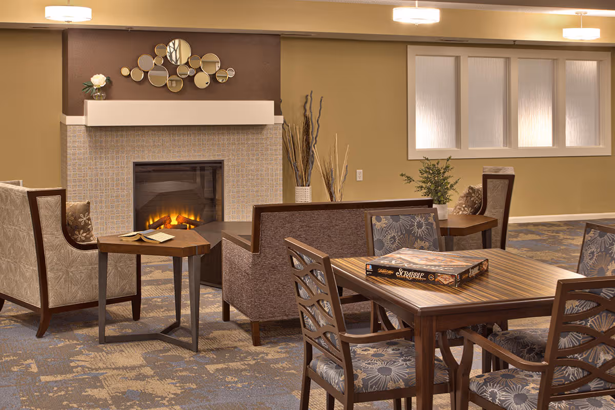 A cozy common area with a lit fireplace, a decorative mirror above the mantle, and comfortable seating including armchairs and a sofa. There is a wooden table with a Scrabble board game on it and another small table with an open book. The room has warm lighting and a carpeted floor with a patterned design.
