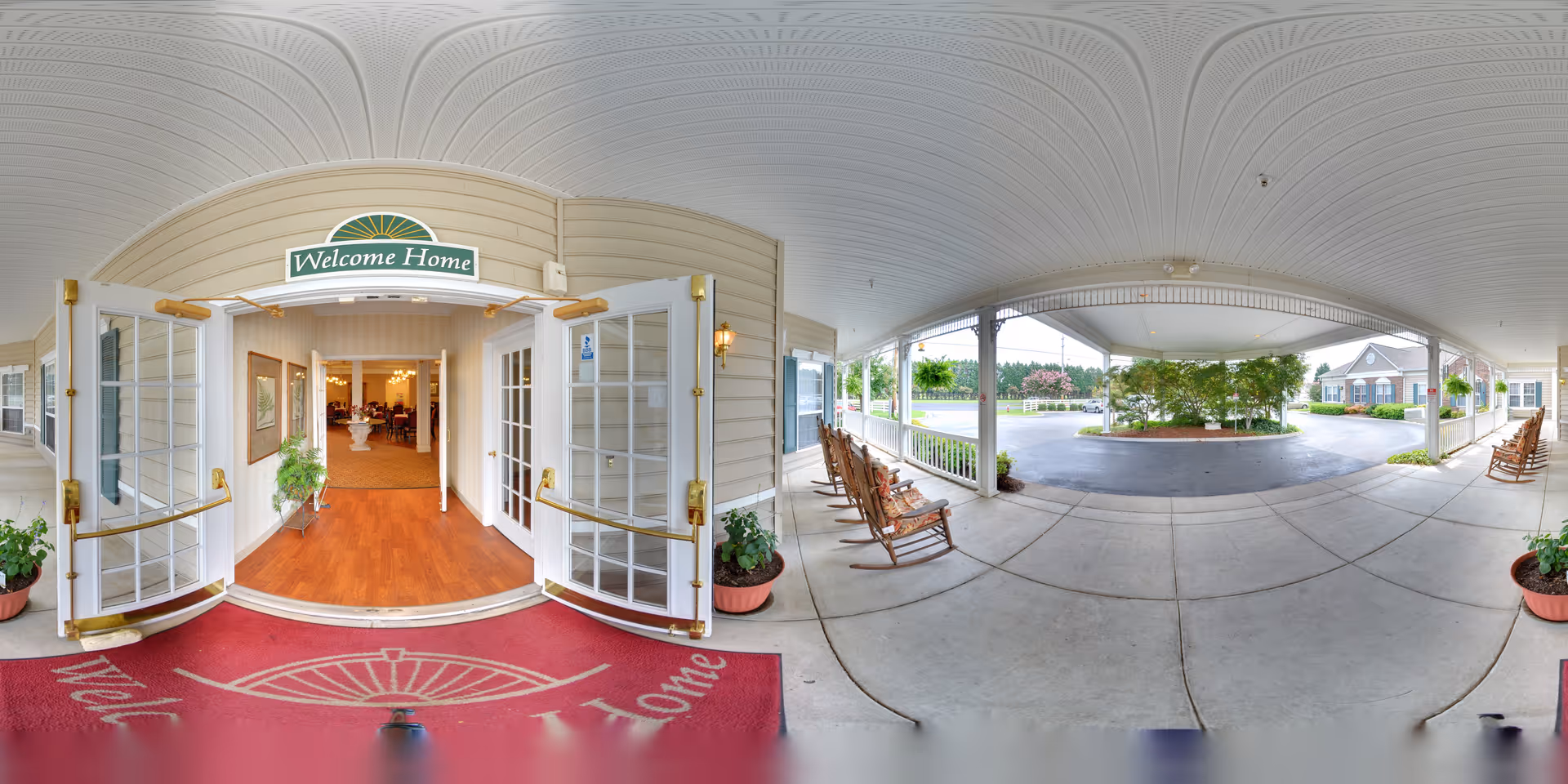 Wide-angle view of the entrance to a senior living facility with double glass doors open, a red welcome mat on the floor, and a green sign above the doors that reads 'Welcome Home'. To the right and left of the entrance are rocking chairs on a covered porch area with potted plants. Inside, a warmly lit interior with wooden flooring and a floral arrangement is visible.