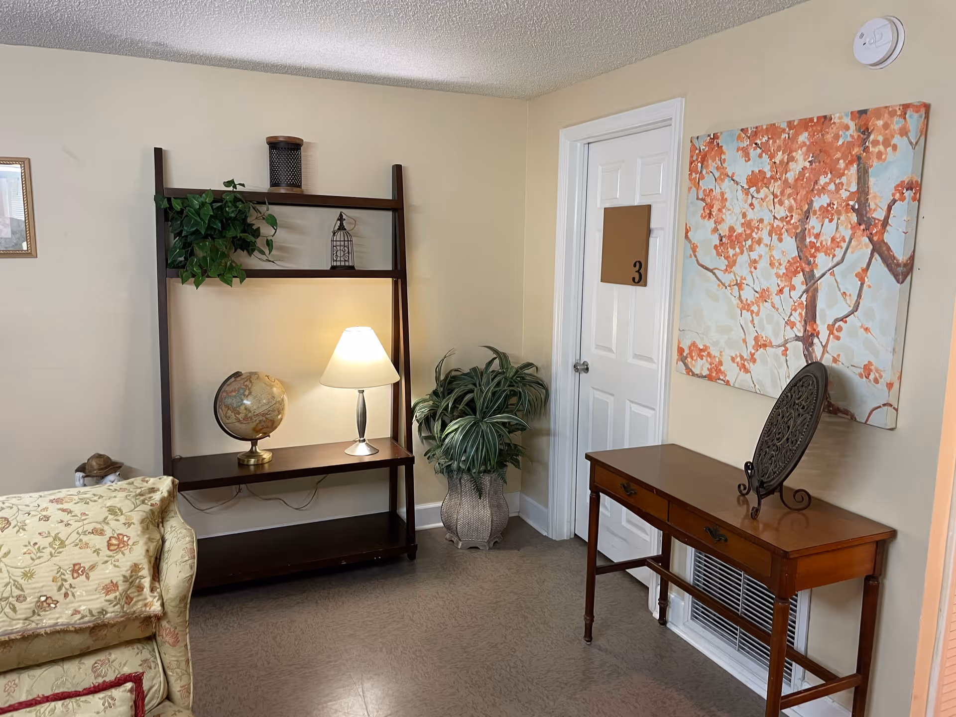 A cozy interior corner of a room featuring a wooden shelf with a globe, a lit table lamp, and a potted plant. Next to the shelf is a wooden console table with two drawers, a decorative plate on a stand, and a large painting of orange blossoms on the wall above it. A white door with the number 3 is visible in the background, and part of a floral-patterned armchair is seen on the left side.