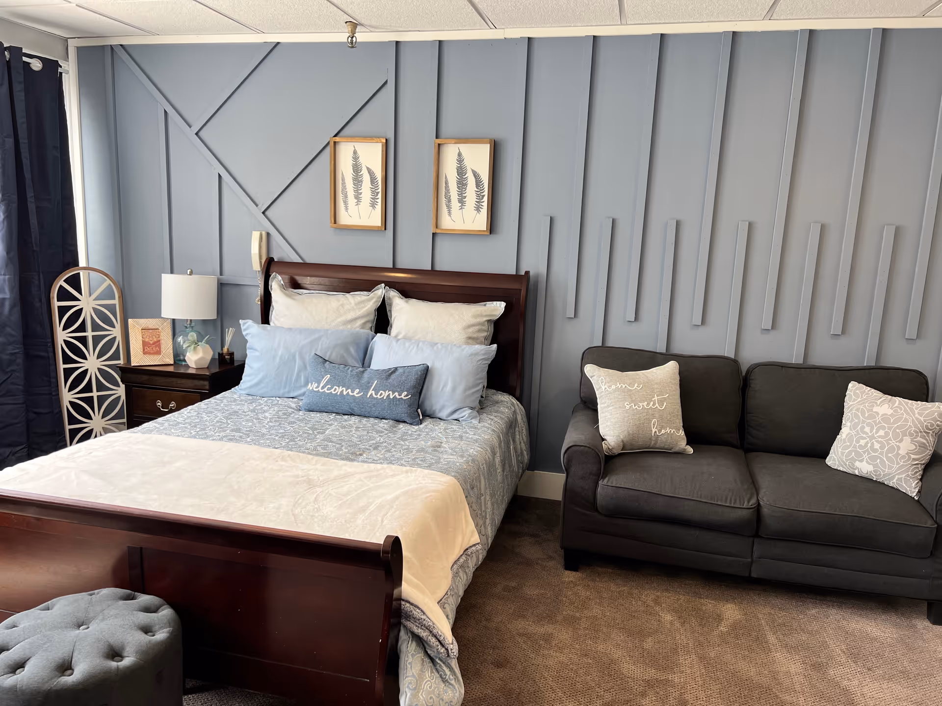 A cozy bedroom with a wooden bed frame, light blue and white bedding, and multiple pillows including one that says 'welcome home'. Next to the bed is a dark wooden nightstand with a lamp and decorative items. On the right side, there is a dark gray sofa with two decorative pillows, one saying 'home sweet home'. The wall behind the bed and sofa is painted light blue with decorative molding and two framed botanical prints. The floor is carpeted in a neutral tone.