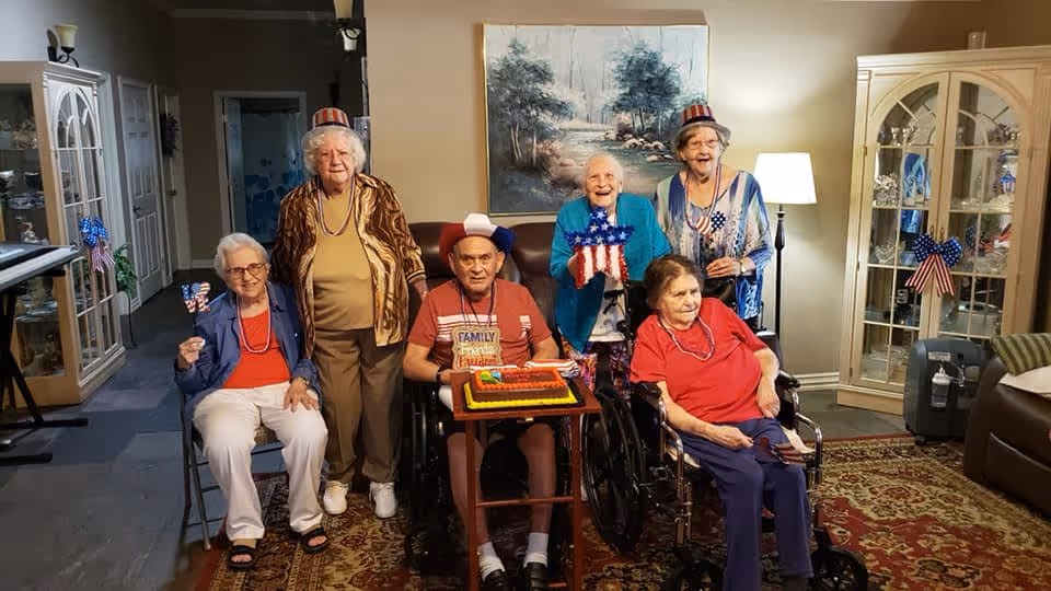 A group of six elderly people, some seated and some standing, celebrating indoors with patriotic decorations and a cake. Two individuals are in wheelchairs, and several are wearing red, white, and blue hats and holding American flag-themed items. The room has a warm, homey atmosphere with a large painting on the wall, display cabinets, and a patterned rug.