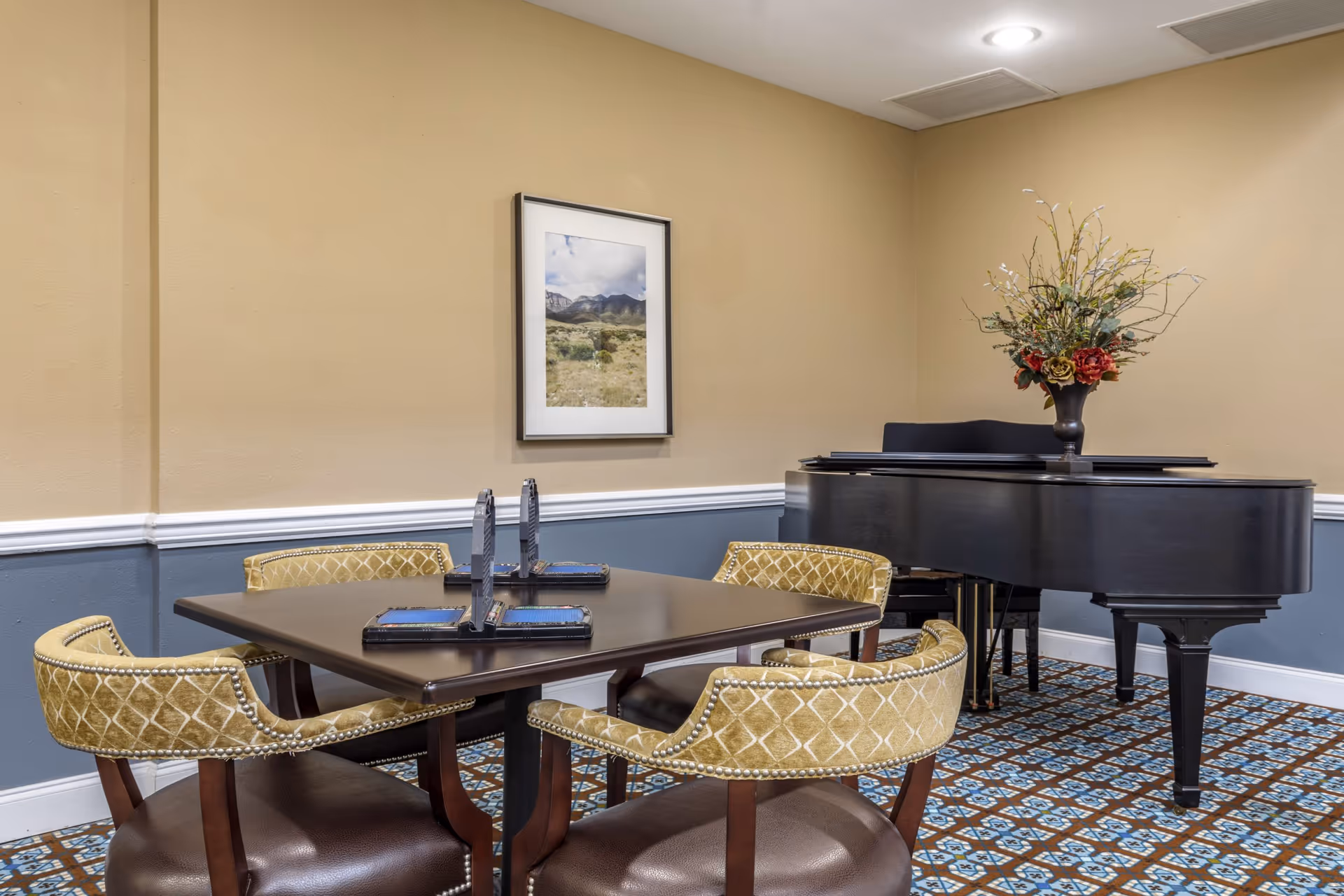 A room with a square dark wood table surrounded by four upholstered chairs with patterned yellow backs and brown seats. On the table are four game boards with racks for tiles, suggesting a game setup. In the background, there is a black grand piano with a floral arrangement on top. The walls are painted beige on the upper half and blue on the lower half, separated by white trim. A framed landscape photograph hangs on the wall above the table. The floor is covered with a patterned carpet in blue, brown, and white tones.