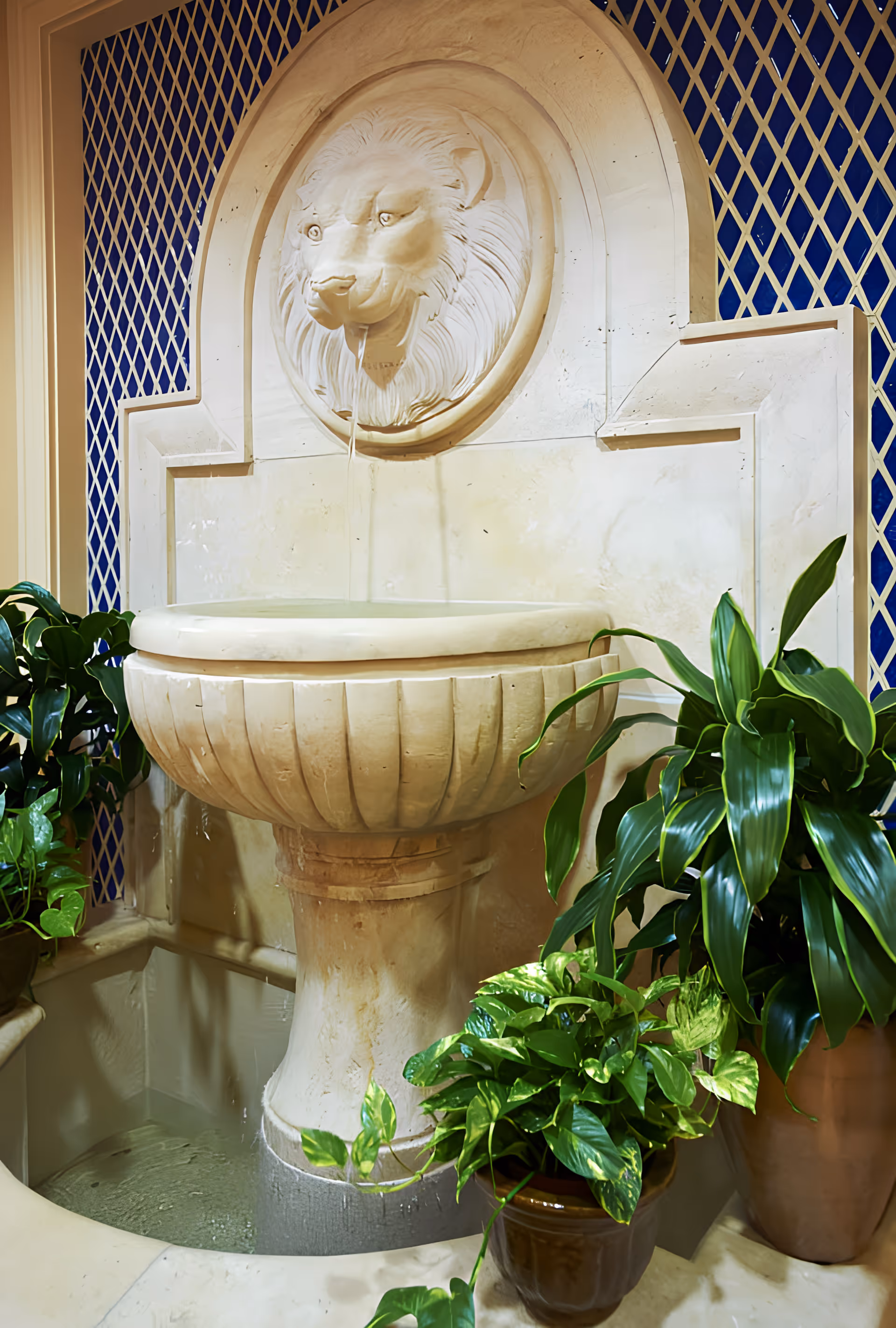 Indoor decorative stone lion-head wall fountain with a basin surrounded by potted green plants.