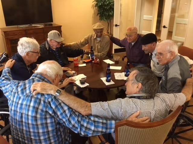 A group of elderly men sitting around a round table in a living room or common area, with their arms around each other's shoulders in a gesture of camaraderie. There are drinks and snacks on the table, and a television and a plant are visible in the background.