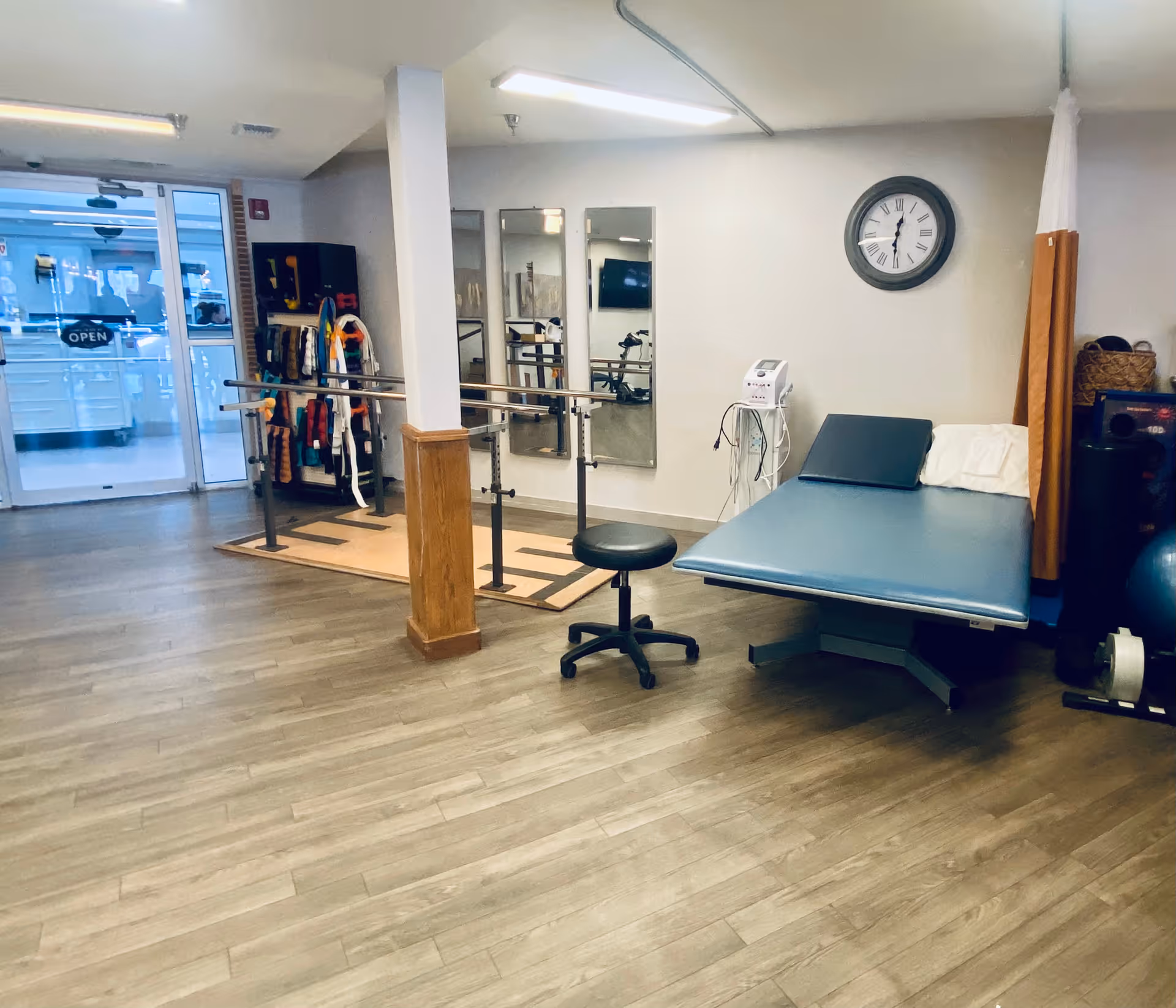 A rehabilitation/therapy room with parallel bars, a treatment table, a rolling stool, mirrors and exercise equipment near an entrance.