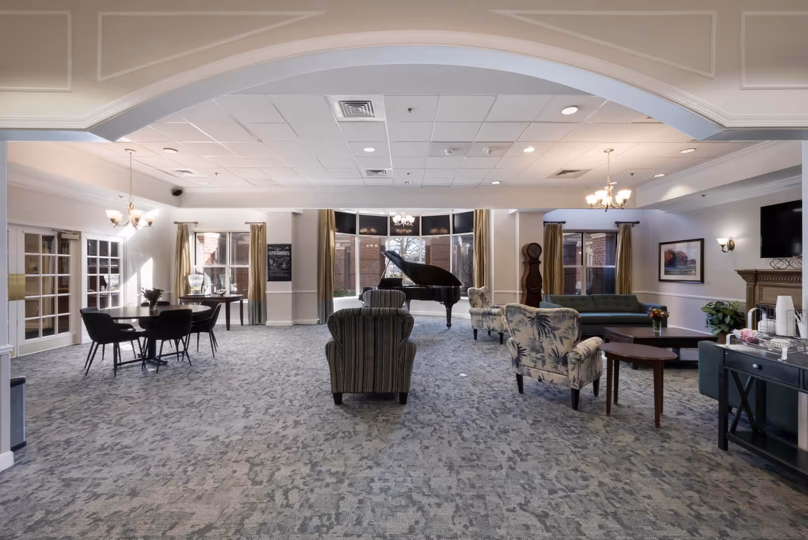 A spacious and well-lit common area in a senior living facility featuring a grand piano near large windows, several upholstered chairs and sofas arranged around coffee tables, chandeliers hanging from the ceiling, and a side table with refreshments.