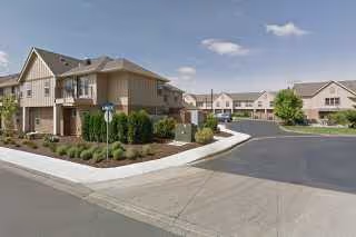 Exterior view of a senior living facility named Maple Valley, showing multiple beige buildings with peaked roofs, landscaped greenery, and a paved driveway under a partly cloudy sky.