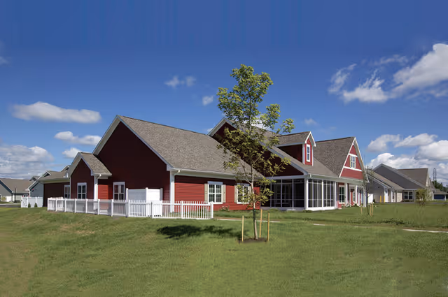 Exterior view of a senior living facility with red buildings, white trim, and a white fence, set against a blue sky with scattered clouds and green grass in the foreground.
