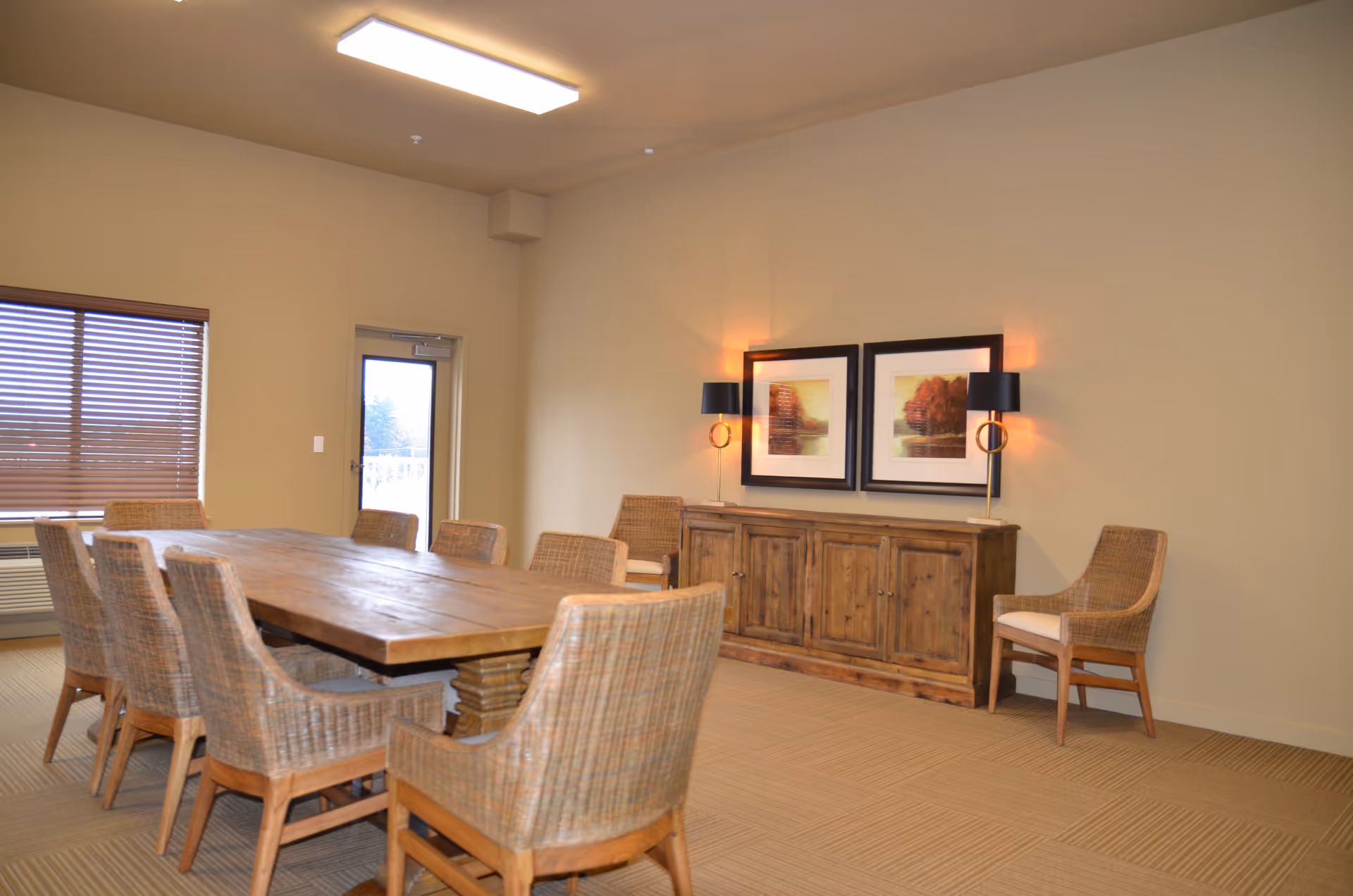 Dining room with a long wooden table surrounded by wicker chairs, a wooden sideboard, framed artwork, and wall lamps.