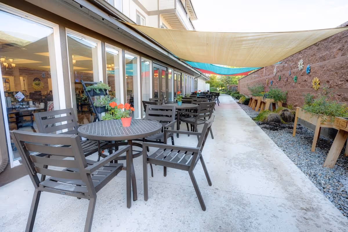 Outdoor patio area with multiple round metal tables and chairs arranged along a concrete walkway. Each table has a small potted plant with flowers. The patio is shaded by large fabric canopies overhead. On the right side, there are raised wooden garden beds with plants and decorative flower wall hangings on a brick wall. Large windows on the left side show an indoor dining area inside the building.