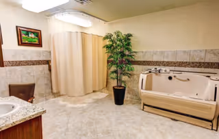 A spacious bathroom with beige tiled floors and walls, featuring a walk-in bathtub with safety handles, a shower area with a beige curtain, a granite countertop with a sink, a brown wicker chair, a potted green plant, and a framed painting on the wall.