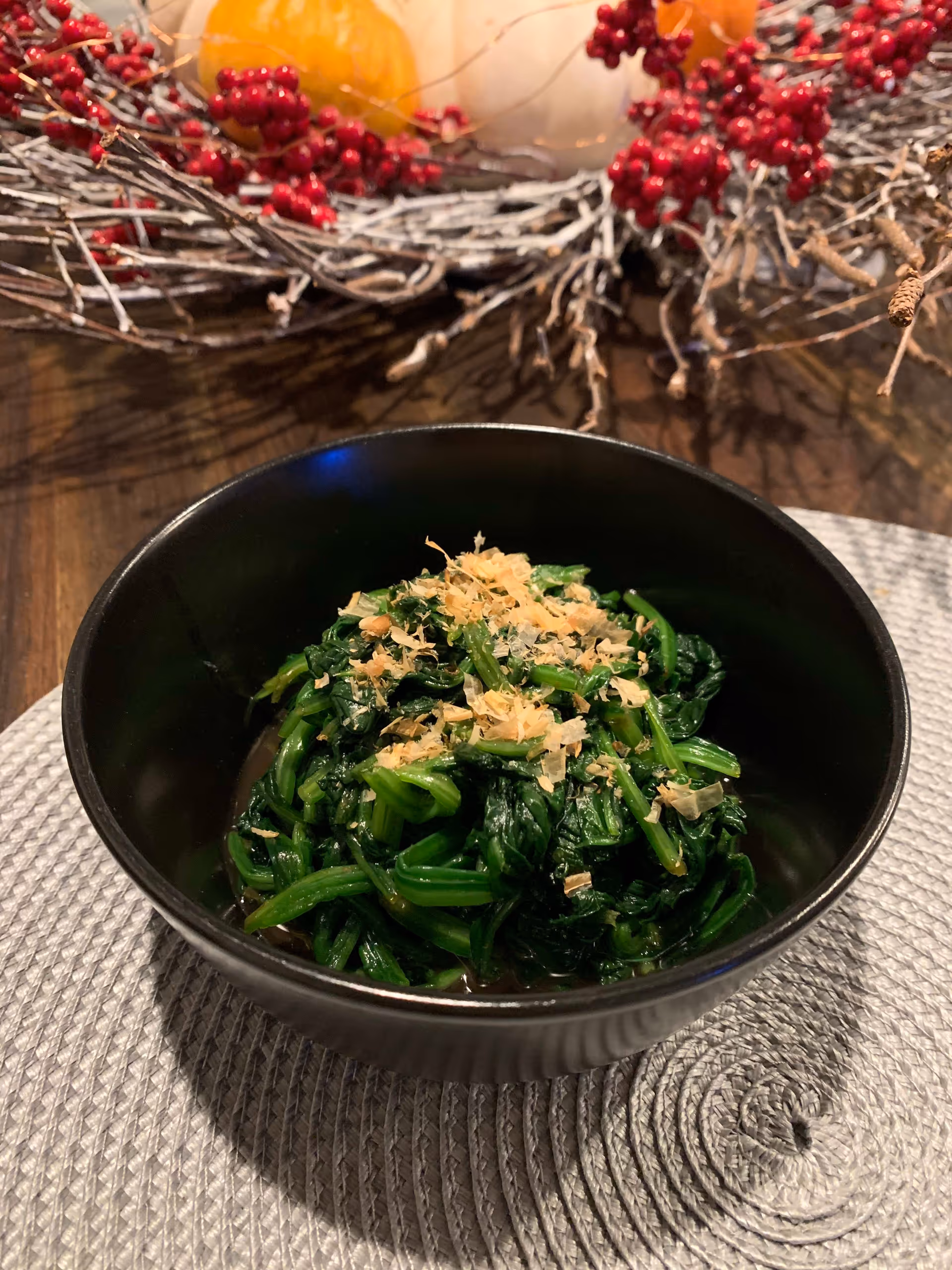 Black bowl of cooked greens topped with flakes on a woven placemat with an autumnal twig and berry centerpiece behind it.