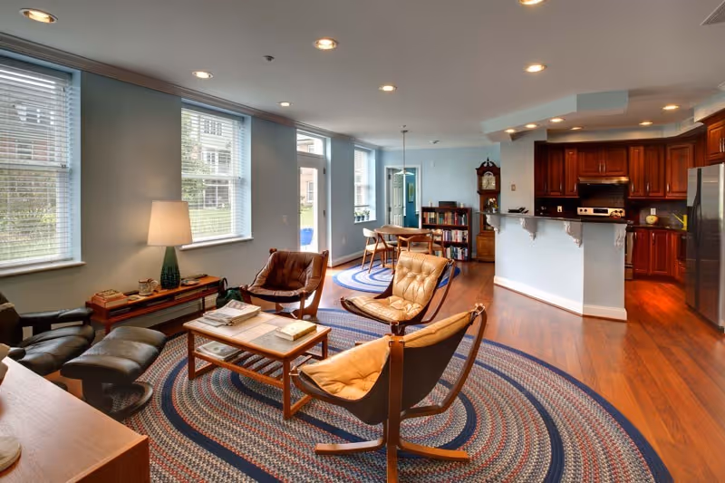Open living room with leather lounge chairs around a coffee table on a round braided rug, large windows, and an open kitchen with wooden cabinets in the background.